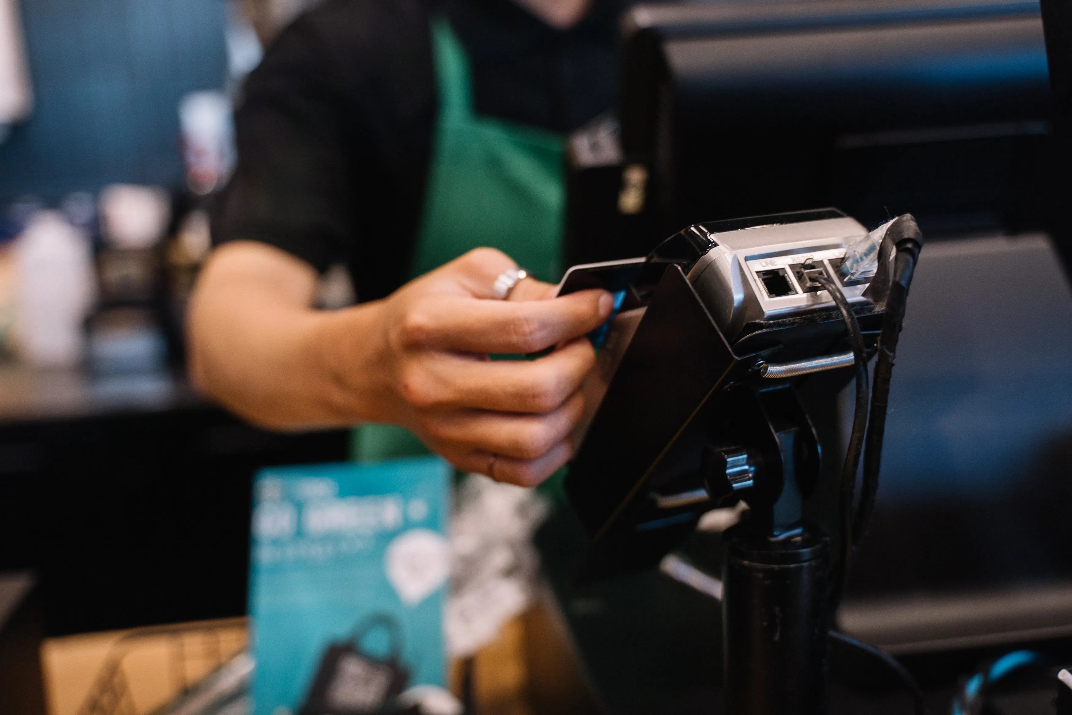 A barista holding a payment terminal while a customer taps a credit card at a busy coffee shop counter