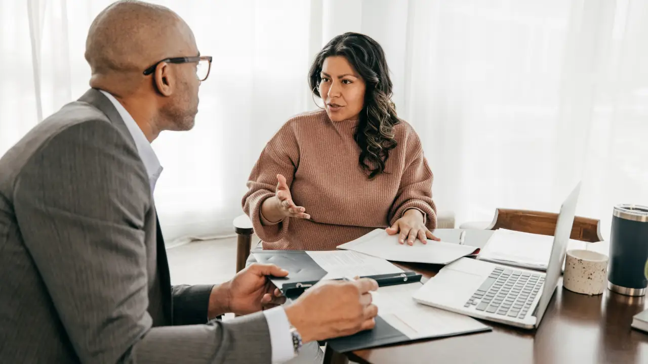 A borrower sitting across from a loan officer in a small office, reviewing a mortgage statement together on paper