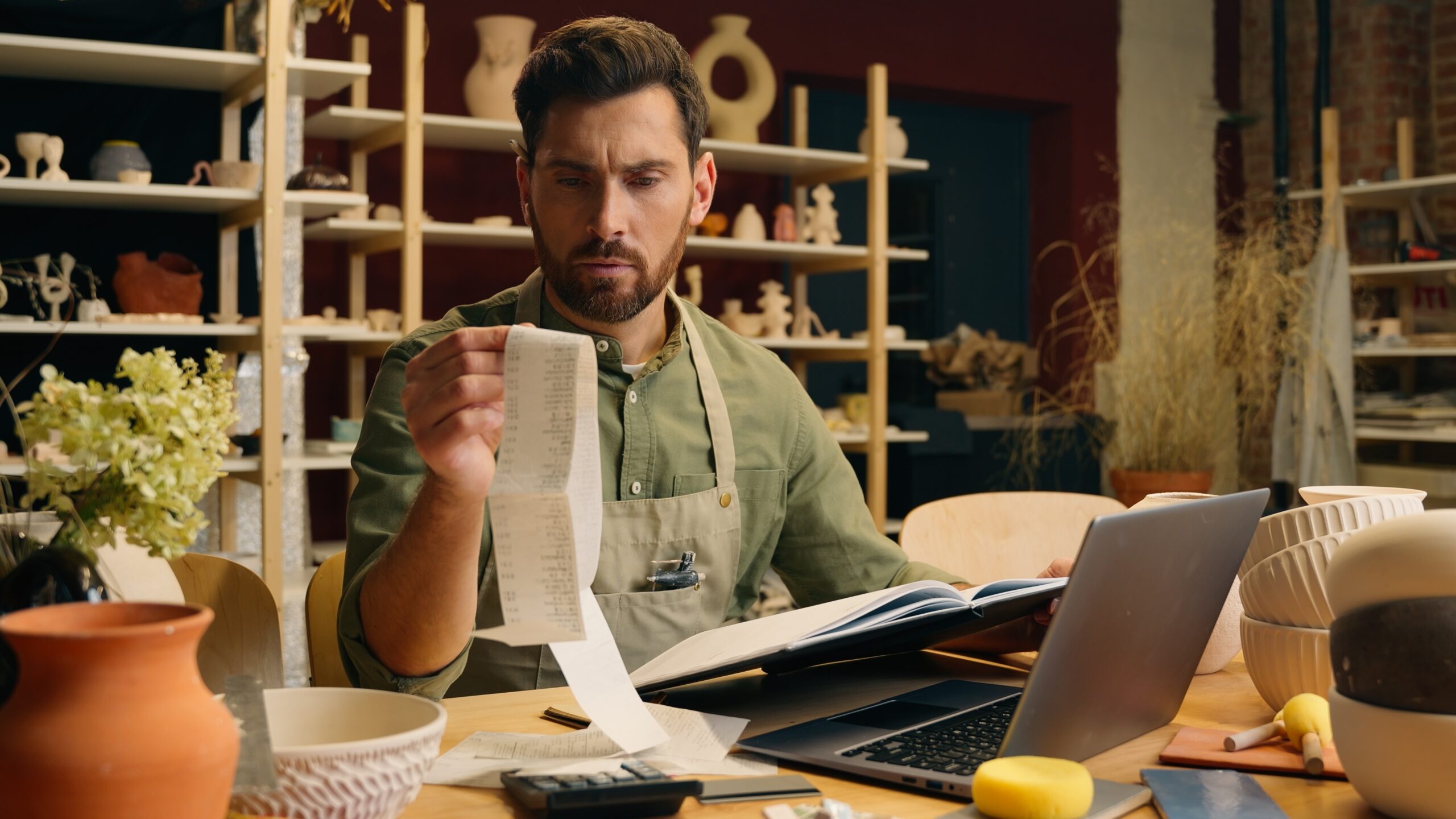 A business owner in a small workspace studying cash flow on a laptop while paper invoices and a calculator sit on the desk