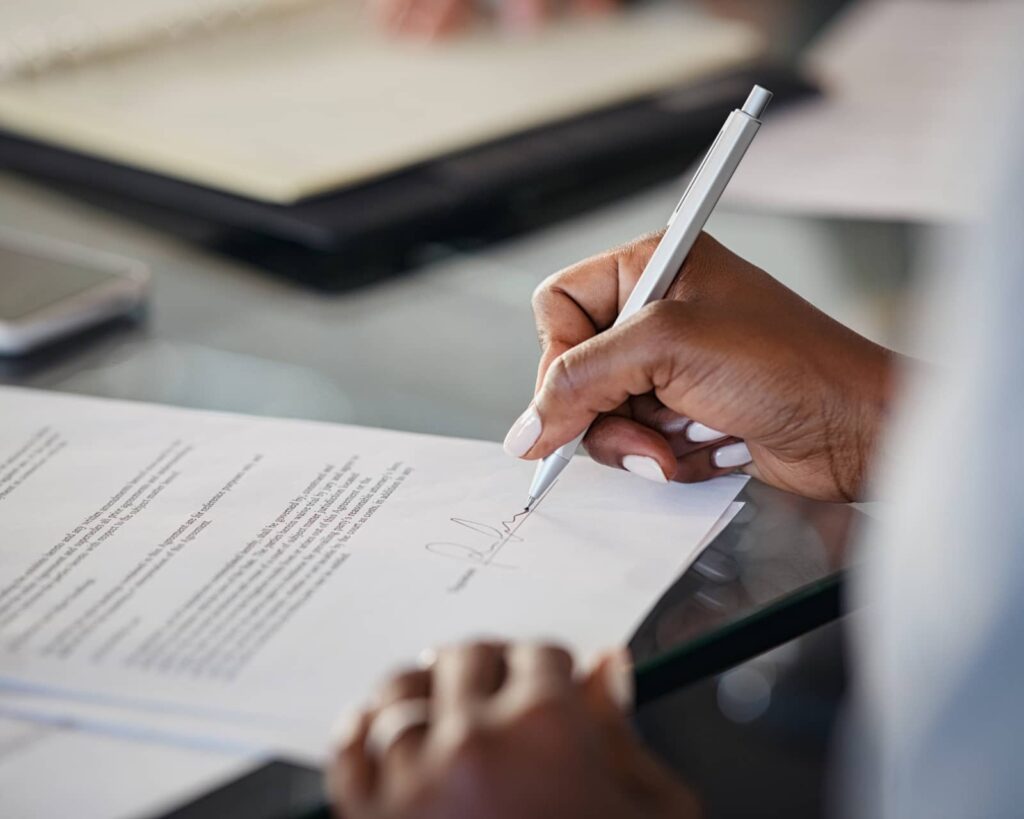 A homebuyer sitting at a kitchen table signing mortgage paperwork with a lender representative nearby, natural indoor light