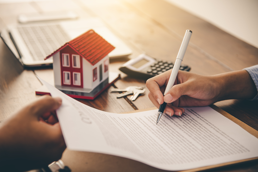 A homeowner seated at a kitchen table signing mortgage paperwork with a lender across from them.