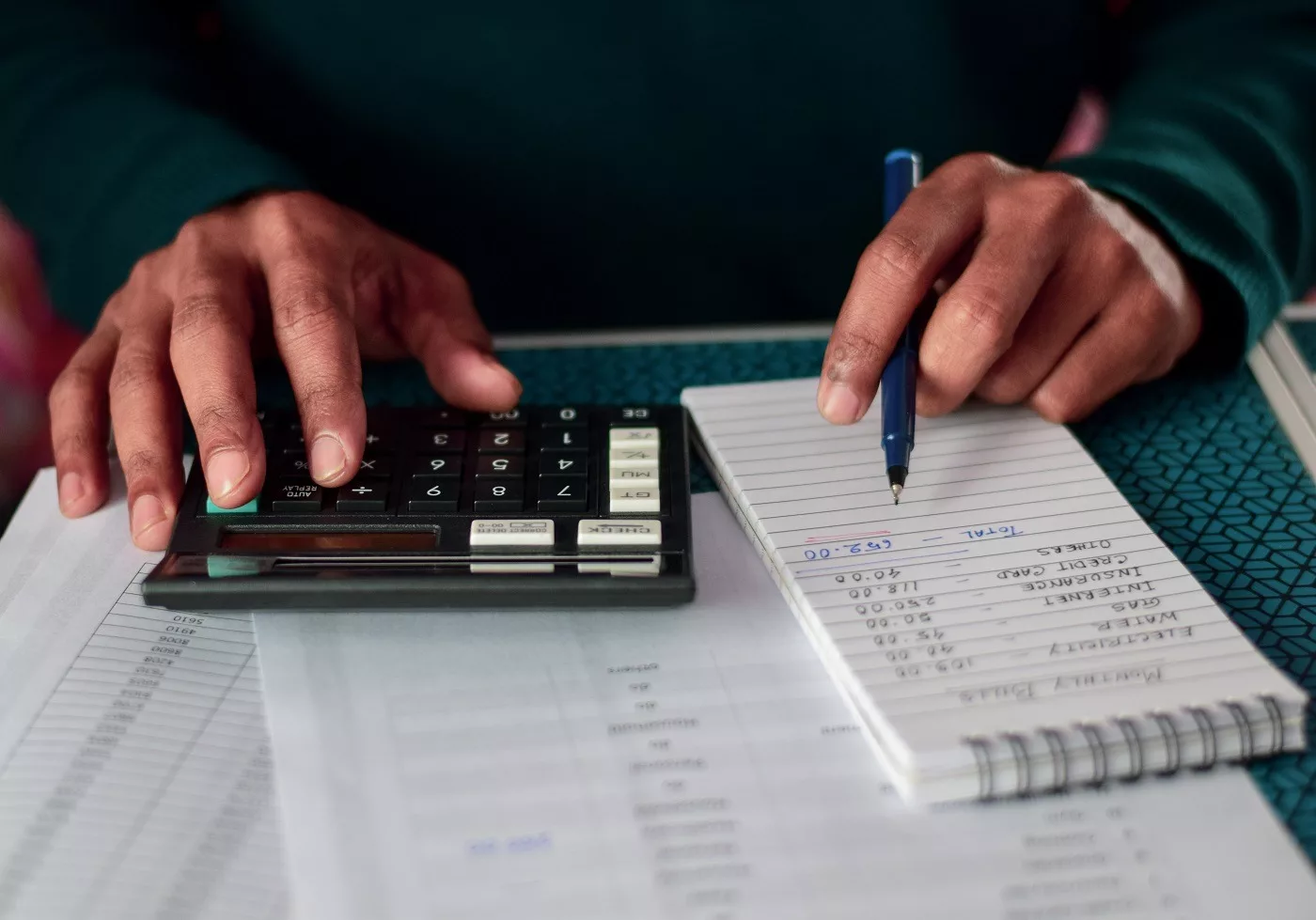 A homeowner sitting at a dining table making monthly bill payments on a laptop with a paper budget sheet beside them