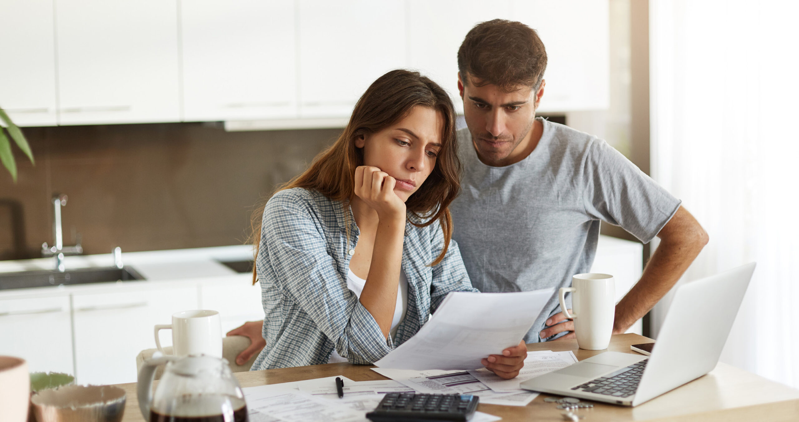 A homeowner sitting at a kitchen table reviewing a mortgage statement and a laptop budget spreadsheet, natural indoor light, candid photo