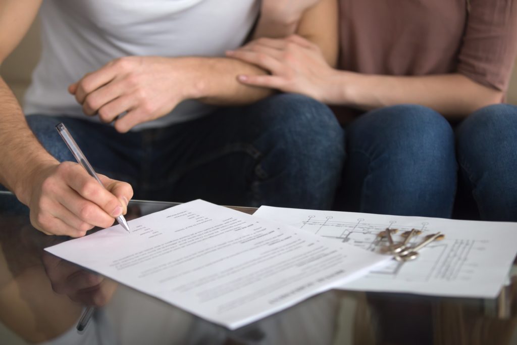 A homeowner sitting at a kitchen table signing mortgage-related paperwork with a pen beside a laptop and a stack of documents