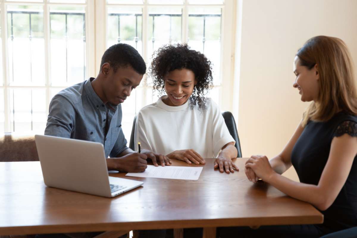 A landlord and tenant seated at a kitchen table in a rental home, reviewing and signing a lease agreement with keys on the table