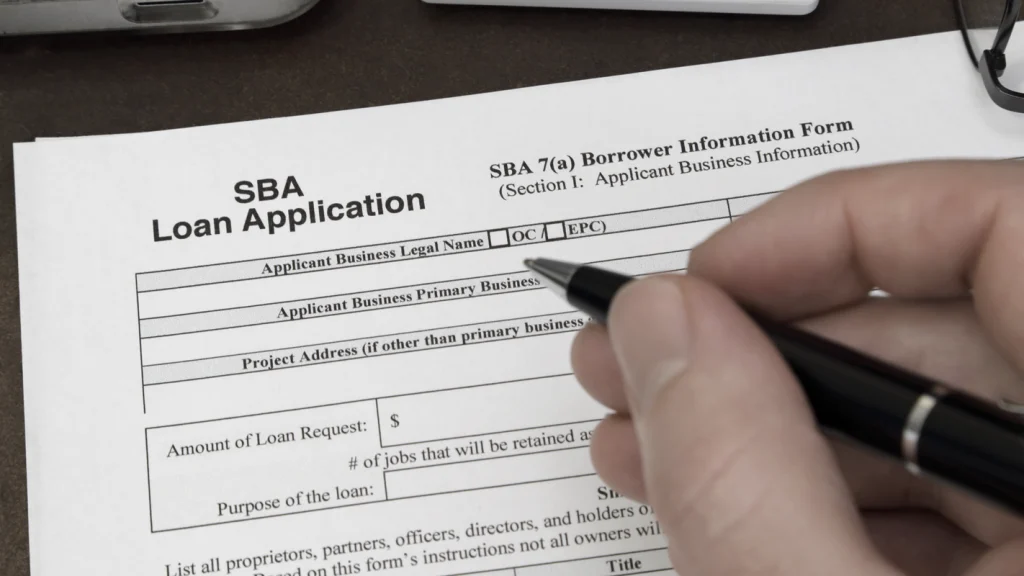 A small business owner sitting at a desk reviewing an SBA loan application packet with a calculator and a laptop.