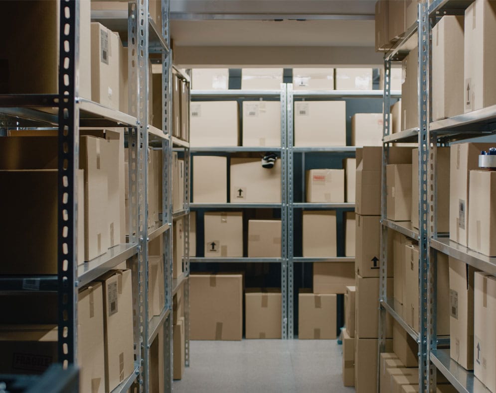 A small retail storeroom with metal shelves holding labeled boxes and products, a shop owner counting inventory with a clipboard
