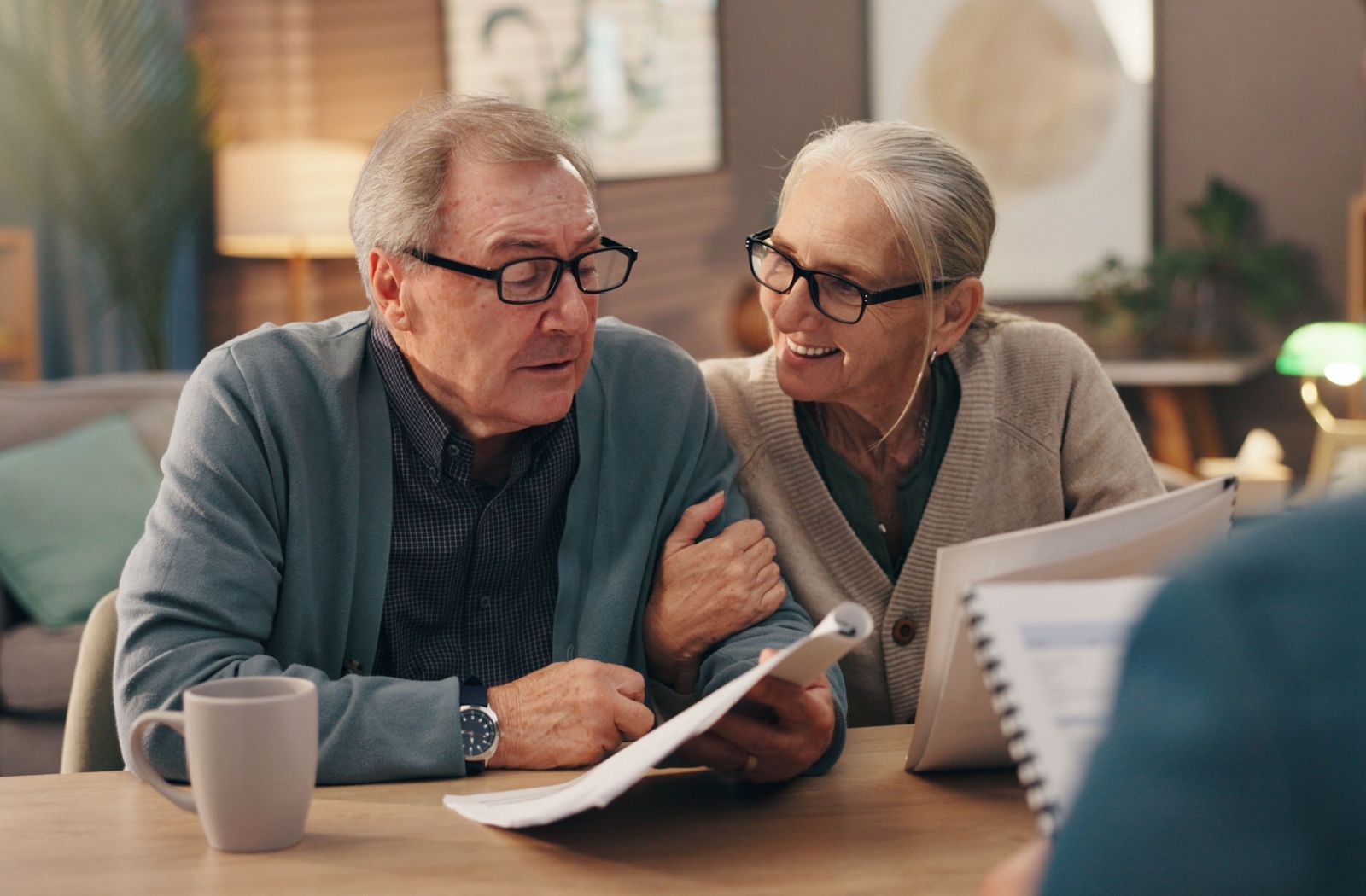 An older couple sitting at a dining room table reviewing retirement account statements with a notebook and reading glasses, warm indoor lighting
